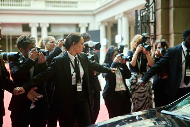 Men and woman in black suits with arms outstretched, bank of cameras in background as photographers capture red carpet event.