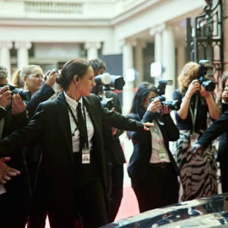 Men and woman in black suits with arms outstretched, bank of cameras in background as photographers capture red carpet event.